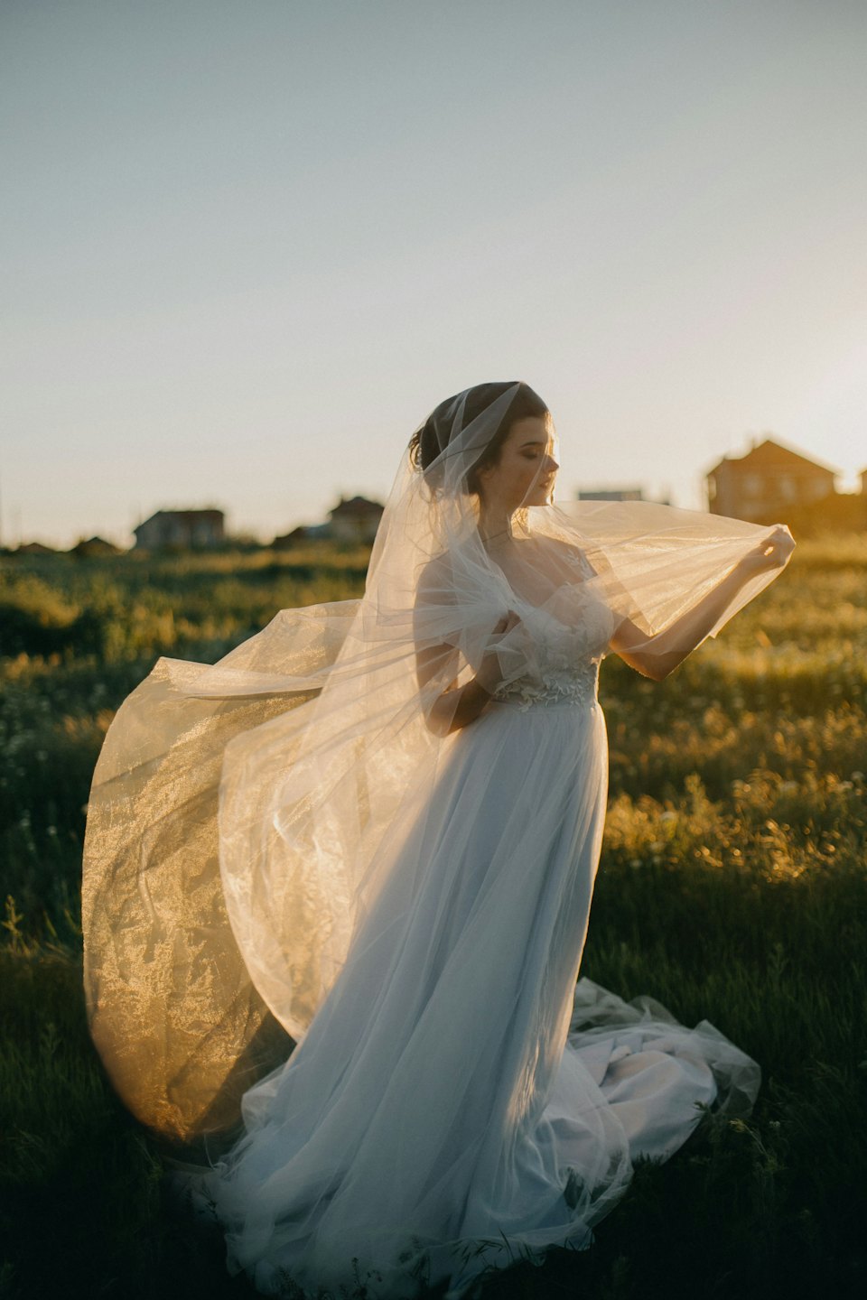 Bride in a white gown holding veil in a green field.